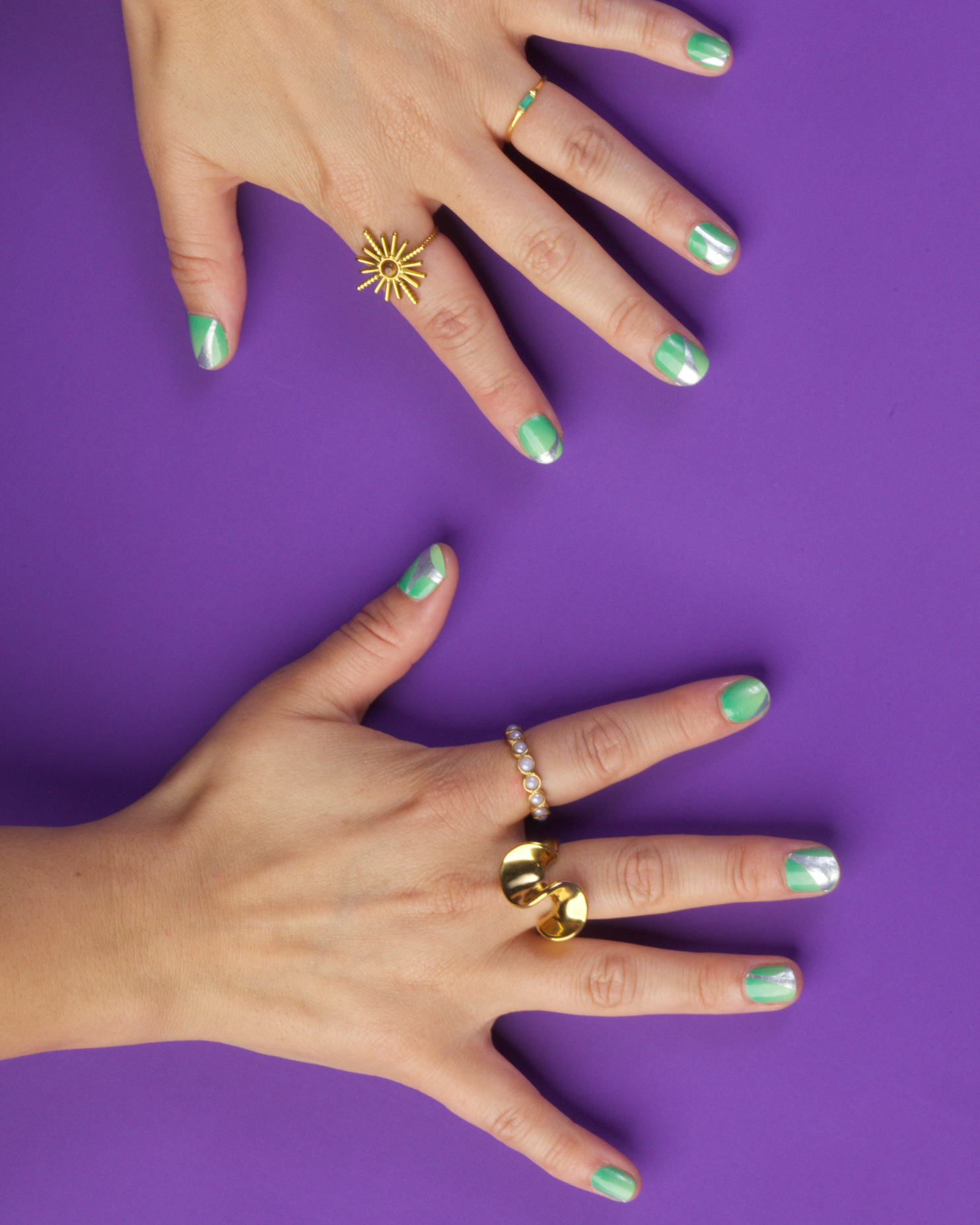 Two hands with green nail wraps and various rings on a purple background